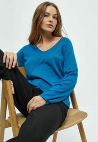 Blauer V-Ausschnitt-Pullover aus weichem Stoff, mit langen Ärmeln. Das Modell sitzt auf einem Holzstuhl und trägt schwarze Hosen.