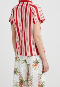 Camisa de rayas rojas y blancas de manga corta con un tejido texturizado; cuello y corte relajado. Llevada sobre pantalones de pierna ancha con estampado floral.