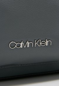 Matériau en cuir gris avec une texture lisse. Logo "Calvin Klein" argenté affiché en évidence à l'avant. Design minimaliste.