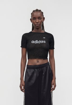 Donna con capelli intrecciati che indossa una maglietta corta nera texturizzata Adidas e pantaloni sportivi neri larghi, in piedi davanti a uno sfondo semplice.