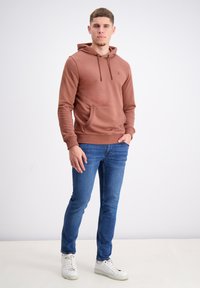 Bruine hoodie met voorzak, verstelbare trekkoorden en geribde manchetten, gecombineerd met blauwe jeans en witte sneakers op een lichte achtergrond.