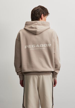 Osoba nosząca beżową bluzę z kapturem z napisem „PEGADOR Certified Authentic Streetwear” na plecach oraz jasne teksturowane spodnie w czarne paski.