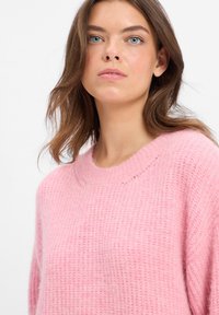 Jonge vrouw met blauwe ogen en bruin haar, gekleed in een zacht roze textuursweater, die licht omhoog kijkt tegen een witte achtergrond.