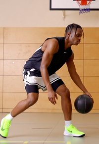 Sort tank top og hvide shorts med sorte accenter. Lyse neongrøn Nike-sko. Atlet, der dribler en basketball i et gymnastikmiljø.