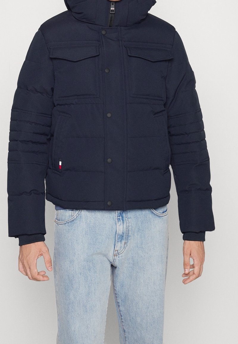 Homme portant une veste matelassée bleu marine avec poches à rabat et un jean bleu clair sur fond uni