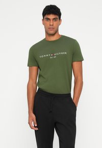 Oliven grønn bomulls T-skjorte med rund hals, med teksten "TOMMY HILFIGER EST. 1985" i hvitt og rødt. Brukt med svarte bukser.