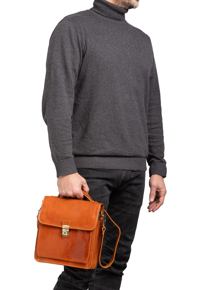 Pochette in pelle marrone con finitura lucida, chiusura metallica e tracolla removibile, tenuta da una persona con un maglione grigio e pantaloni neri.