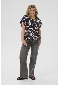 Femme souriante aux cheveux blonds portant une chemise à motifs noir et blanc, un jean gris et des sandales noires, se tenant devant un fond uni.