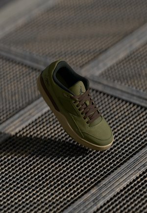 Buty sportowe oliwkowe z brązowymi sznurówkami i beżową podeszwą, sfotografowane unoszące się nad teksturowanym metalowym rusztem.