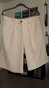 Beige Shorts aus Baumwolle mit flachem Frontdesign, einem Knopfverschluss und zwei seitlichen Taschen. Glatte Textur und knielange Ausführung.