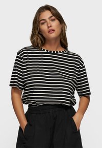 Mujer con una camiseta de rayas horizontales en blanco y negro de mangas cortas y pantalones negros con cintura elástica, con las manos en los bolsillos.