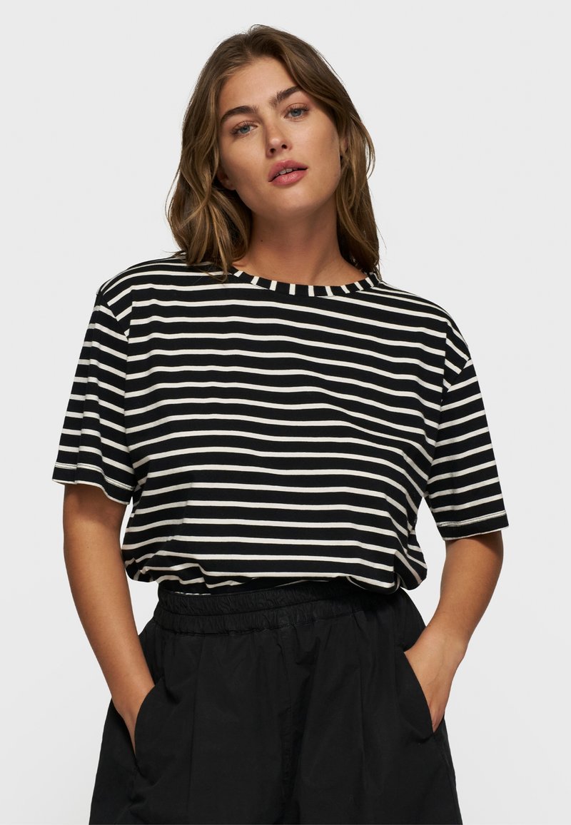 Mujer con una camiseta de rayas horizontales en blanco y negro de mangas cortas y pantalones negros con cintura elástica, con las manos en los bolsillos.
