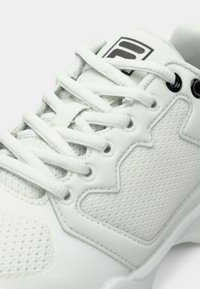 Baltie sporta kedas ar baltām šņorēm, acs un sintētiskajiem paneļiem, un melnu logo uz mēles, attēlots tuvplānā.