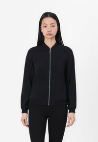 Veste bomber noire avec une fermeture éclair, des poignets côtelés, une poche frontale et une texture lisse. Le modèle la porte sur un pantalon noir.