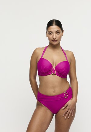 Vrouw met donker haar in een roze tweedelige badpak met gouden gespaccenten, staand tegen een effen witte achtergrond.