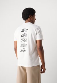 Witte katoenen T-shirt met zwarte tekst en logo's op de achterkant. Korte mouwen, ronde hals, standaard pasvorm. Glad textuur.