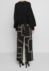 Svart ribbad tröja med voluminösa ärmar, i kombination med vida byxor med en beige geometrisk mönstring på svart bakgrund.