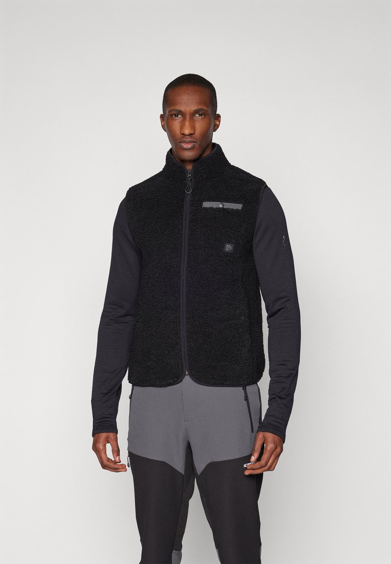 Μαύρο γιλέκο από φλις με ψηλό γιακά και μπροστινό φερμουάρ, διαθέτοντας μια γκρίζα αξεσουάρ τσ�έπη και υφή υφάσματος, φορεμένο πάνω από μαύρη μπλούζα με μακριά μανίκια.