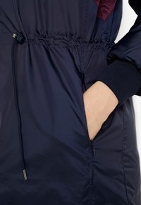 Chaqueta acolchada azul marino con cintura y puños elásticos. Cuenta con un cierre de cordón y un bolsillo con cremallera en el lado.