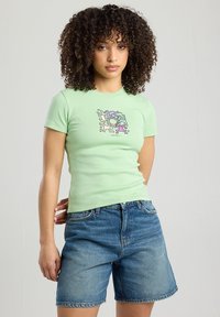 T-shirt ajusté vert menthe, à manches courtes, avec un imprimé graphique en violet et noir au centre. Assorti avec un short en denim.