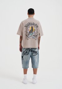 Beige shirt met korte mouwen met grafische print op de rug; denim shorts met zwarte letters en grafische details; witte sneakers en enkelkousen.