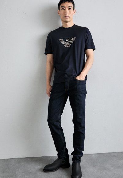 Camiseta navy con un diseño de logo en un color claro, combinada con jeans de mezclilla oscuros. El modelo lleva botas negras hasta el tobillo. Estilo sencillo y moderno.