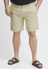 Beige bomullsshorts med standard passform, sidelommer og knappelukking. Brukt med brune sandaler med dobbel spenne-design.