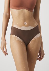 Brune høy midje bikini-truser har en glatt tekstur og en buet livlinjedesign. Modellen viser hud med lyse og mørke flekker.