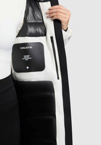 Lang sort og hvid oppustet frakke med en skinnende sort inderside, udstyret med en firkantet lomme mærket "KHUJOTM." Glat tekstur, let vægt.