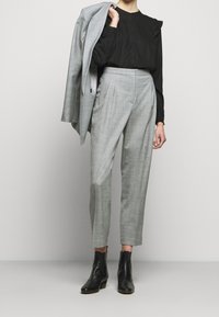 Pantalon à carreaux gris coupe ajustée avec des plis, associé à une chemise noire à manches longues et un blazer gris clair posé sur un bras. Bottines noires.