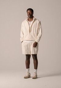 Világosbézs cipzáros dzseki rugalmas mandzsettákkal, összeillő rövidnadrággal párosítva. Fehér póló fölött viselve, bézs tornacipővel és fehér zoknival kiegészítve.