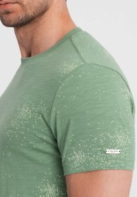 Vihreä t-paita, jossa on teksturoitu pinta, ja jossa on satunnaisesti aseteltu printtikuosi sekä pieni metallinen logo-aksentti helman lähellä. Lyhyet hihat.