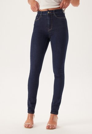 High-waisted skinny jeans in donkerblauwe denim met een gladde textuur, contrasterende stiksels en een vijf-pocket ontwerp. Transparante sandalen zichtbaar.