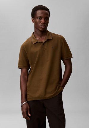 Junger Mann, der ein braunes Poloshirt und schwarze Hosen trägt, steht mit einer Hand in der Tasche vor einem schlichten grauen Hintergrund.