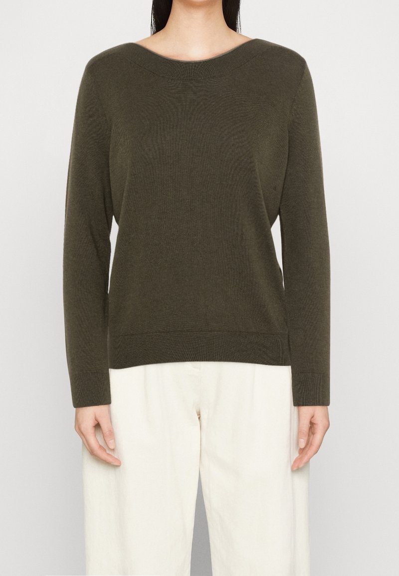 Frau in einem olivgrünen langärmeligen Rundhalspulli und lockeren weißen Hosen, die vor einem schlichten hellen Hintergrund steht.