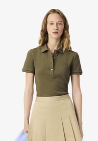 Polo shirt verde oliva con colletto, dotato di tre bottoni e un piccolo logo. Abbinato a una gonna plissettata beige.