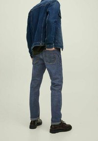 Chaqueta de mezclilla azul oscuro con un diseño clásico, llevada sobre una camiseta negra, combinada con jeans azules ligeramente desgastados y zapatos de cuero negro.