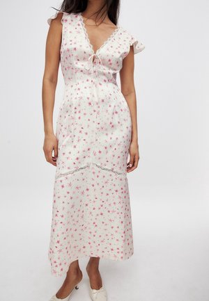 Femme portant une robe midi blanche sans manches avec un imprimé floral rose, un galon en dentelle, un lien à l'avant et des chaussures blanches à talons bas, debout sur fond blanc.