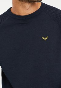 Donkerblauwe trui van breiwerk, met een geribde kraag en een klein geborduurd logo in het geel op de borst.