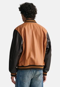 Hombre con cabello rizado, vistiendo una chaqueta de varsity de cuero marrón y negro y vaqueros de denim azul, de espaldas frente a un fondo blanco.