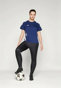 Navy athletic shirt with white accents, paired with black fitted leggings and black soccer shoes, standing on a black-and-white soccer ball.