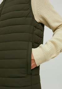 Olivgrüne Steppweste mit horizontalem Quiltmuster und seitlichen Taschen. Über einem beigen Strickpullover mit gerippten Bündchen getragen.