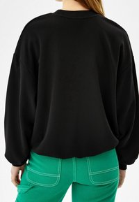 Person som bär en svart långärmad topp och gröna byxor med vit söm, visad bakifrån mot en enkel bakgrund.