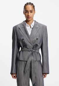 Veste croisée grise à rayures avec une taille ceinturée, des revers crantés et des manches longues, associée à une chemise blanche boutonnée en dessous.