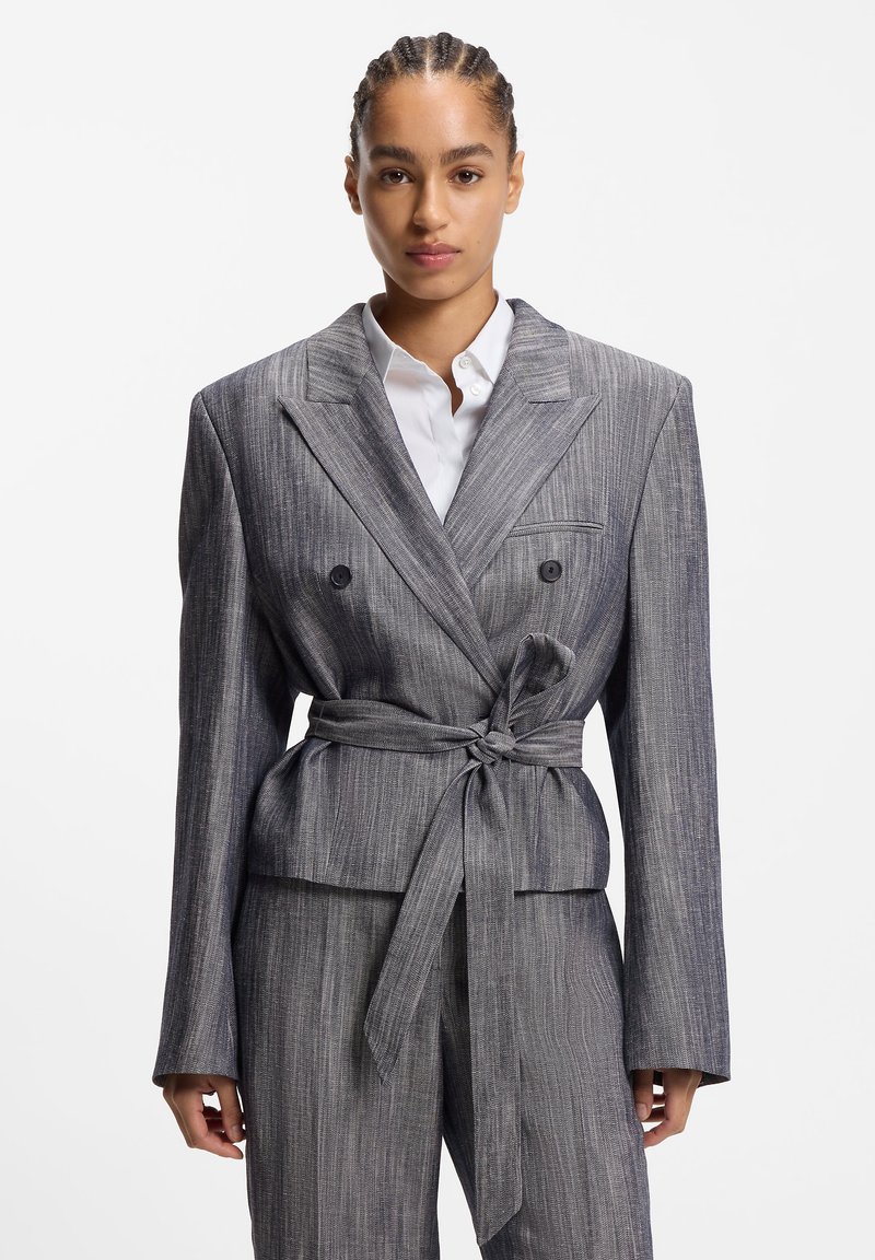 Veste croisée grise à rayures avec une taille ceinturée, des revers crantés et des manches longues, associée à une chemise blanche boutonnée en dessous.