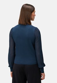 Mørkeblå sweater med lange, stribede gennemsigtige ærmer. Tætsiddende krop, ribbede manchetter og rund hals. Glat tekstur på hoveddelen.