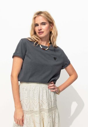 T-shirt en coton gris foncé avec des manches roulées, ornée d'un petit détail en forme de cœur noir ; associé à une jupe de couleur crème avec des motifs en œillets.