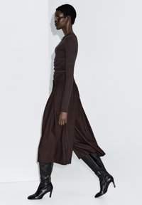 Bruin shirt met lange mouwen gecombineerd met een donkergroene midi-rok en zwarte kniehoge laarzen met hak. Zonnebril maakt de look af.