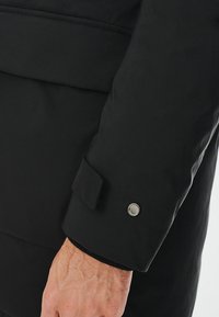 Czarna rękawica kurtki wykonana z gładkiego materiału z akcentem guzika przy mankiecie oraz lekko wyprofilowanym krojem, ukazująca czystą, nowoczesną sylwetkę.