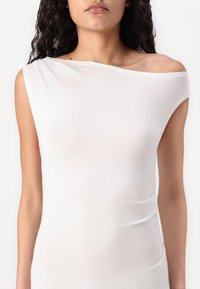 Haut blanc à épaule dénudée en tissu léger, présentant un design ajusté et une texture lisse, avec un léger effet drapé sur un côté.